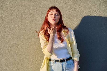 Happy beautiful young hipster female with red hair facial piercing looking at camera making air kiss gesture with hand, sunny gray wall background. Beauty, youth, fashion, joy, happiness, lifestyle