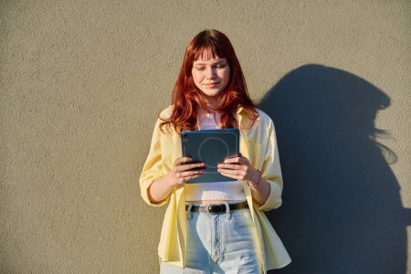 Young attractive female hipster college student using digital tablet outdoor, sunny gray wall background. Education, technology, training, 19,20 years age youth lifestyle concept