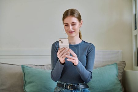 Photo for Happy smiling female teenager using smartphone, sitting on couch at home, girl 16,17, 18 years old texting reading messages. Modern digital technologies for communication, leisure, learning, shopping - Royalty Free Image