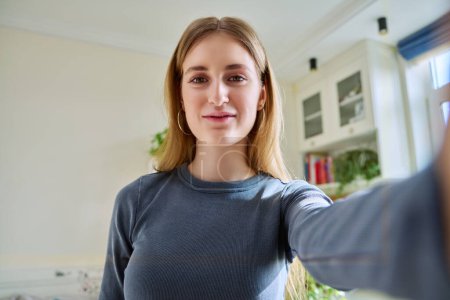 Photo for Close-up selfie portrait of teenage cheerful grimacing young female looking at web camera. Happy teenage girl 16, 17, 18 years old with blond hair, in home interior. Beauty, lifestyle, youth concept - Royalty Free Image