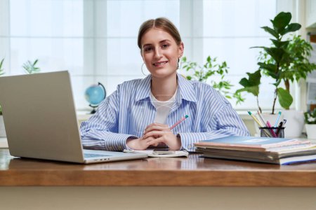 Téléchargez les photos : Portrait d'une lycéenne, jeune étudiante souriante assise au bureau avec un ordinateur portable regardant la caméra. Éducation, formation, apprentissage en ligne, 16, 17, 18 ans - en image libre de droit