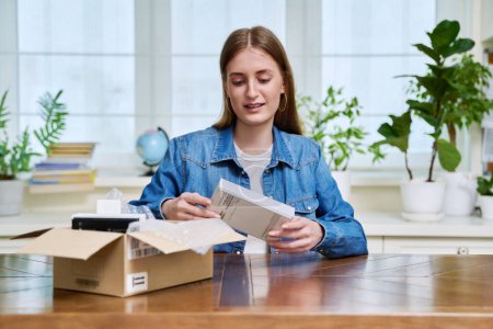 Satisfied young teen female shopper consumer sits at home and unpacks cardboard box with online purchases. Teenager girl unpacking box with new smartphone, headphones. Delivery by mail, online store
