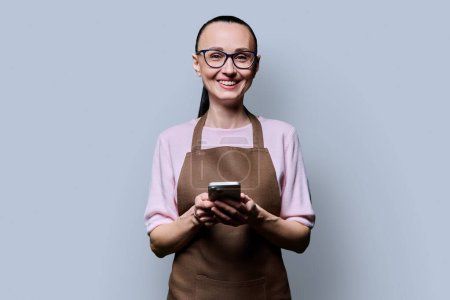 Photo for Portrait of 30s woman in apron with smartphone looking at camera on grey background. Smiling female using mobile phone texting receiving sending order. Technologies applications service small business - Royalty Free Image