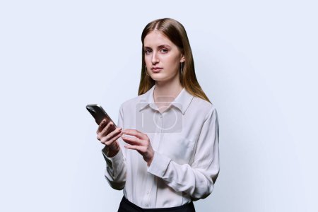 Photo for Teenage student girl 16, 17, 18 years old in white shirt holding smartphone in hands, serious looking at camera on white studio background. Education, technology, high school, college, lifestyle - Royalty Free Image
