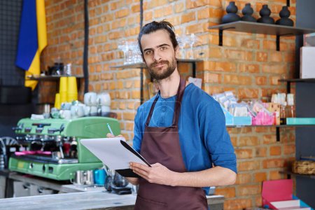 Photo for Young man in apron, food service worker, small business owner entrepreneur with work papers looking at camera near counter of coffee shop cafe cafeteria. Staff, occupation, successful business, work - Royalty Free Image