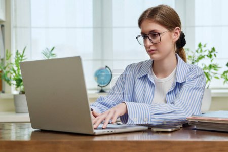 Photo for Young smiling girl typing on laptop computer at home. Female freelancer working remotely, high school, college student studying online preparing for exams. Technology youth training education work - Royalty Free Image