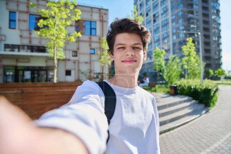 Téléchargez les photos : Selfie portrait de jeune homme heureux beau avec les cheveux bouclés, gars université étudiant en plein air. Jeune, style urbain, 19,20 ans homme, concept lifestyle - en image libre de droit