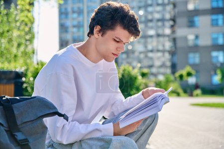 Téléchargez les photos : Beau jeune étudiant universitaire masculin lisant un livre assis sur des marches extérieures. Éducation, connaissance, jeunesse 19-20 ans, passe-temps littéraire et loisirs - en image libre de droit