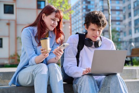 Photo for Teenage college students guy and girl talking, using laptop smartphone, sitting outdoor near educational building. Youth 19-20 years old, education, technologies, lifestyle, friendship concept - Royalty Free Image