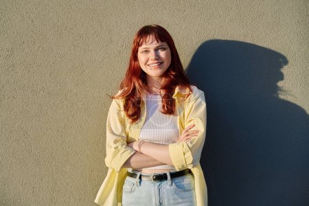 Outdoor portrait of happy confident young hipster female with red hair facial piercing looking at camera with crossed arms sunny gray wall background. Beauty, youth, fashion, joy, happiness, lifestyle