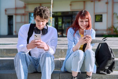 Teenage youth guy and girl university college students sitting outdoor on campus steps using smartphone. Technology, friendship, lifestyle concept