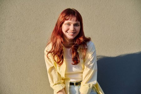 Outdoor portrait of happy beautiful young hipster female with red hair facial piercing looking at camera against sunny gray wall background. Beauty, youth, fashion, joy, happiness, lifestyle concept