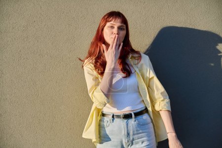 Happy beautiful young hipster female with red hair facial piercing looking at camera making air kiss gesture with hand, sunny gray wall background. Beauty, youth, fashion, joy, happiness, lifestyle