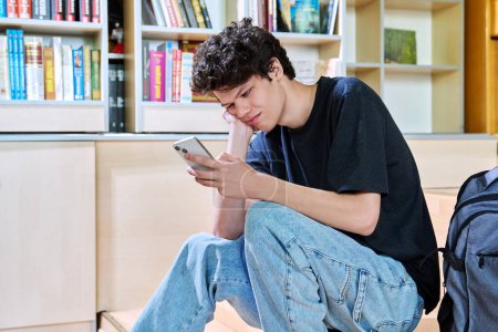 Triste molesto joven estudiante universitario masculino mirando el teléfono inteligente mientras está sentado en el aula. Emociones negativas, dificultades, problemas, concepto de juventud