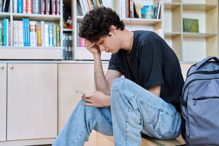 Triste molesto joven estudiante universitario masculino mirando el teléfono inteligente mientras está sentado en el aula. Emociones negativas, dificultades, problemas, concepto de juventud