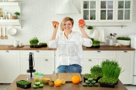 A young beautiful woman is cutting fruit and laughing in the kitchen on the table for a vitamin smoothie made of micro-greens and fruits.