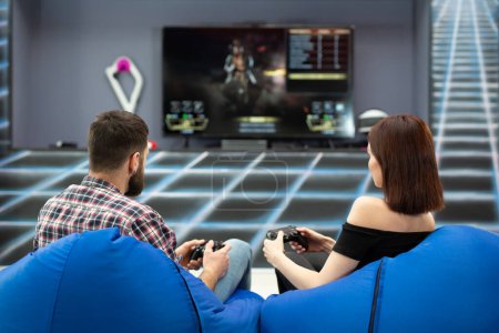 Young couple playing video games, sitting on chairs in a gaming club with controllers in their hands, a rear view from the TV screen.