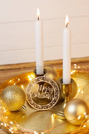 Foto de Velas en candelabros de bronce, decoración de bolas brillantes para árbol de Navidad y placa de madera con letras de Feliz Navidad en un soporte de oro. Fondo de madera blanca. Luces de velas y linternas led - Imagen libre de derechos