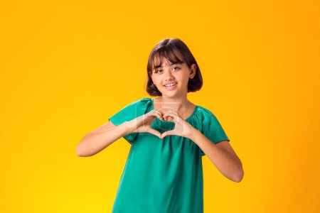 Photo for Smiling kid girl showing heart gesture over yellow background. Love concept - Royalty Free Image
