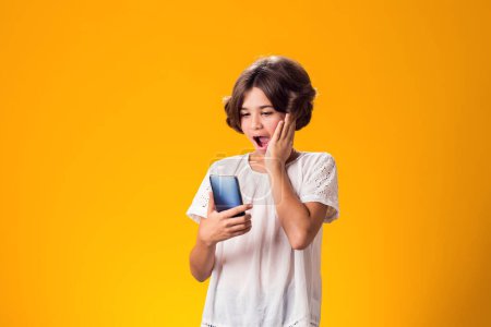 Photo for Surprised kid girl holding smartphone in hand over yellow background - Royalty Free Image