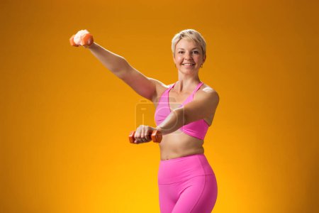Foto de Mujer fitness con pelo corto y rubio sosteniendo pesas aisladas sobre fondo amarillo. Concepto de actividad y asistencia sanitaria - Imagen libre de derechos