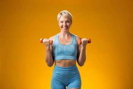 Foto de Mujer fitness con pelo corto y rubio sosteniendo pesas aisladas sobre fondo amarillo. Concepto de actividad y asistencia sanitaria - Imagen libre de derechos