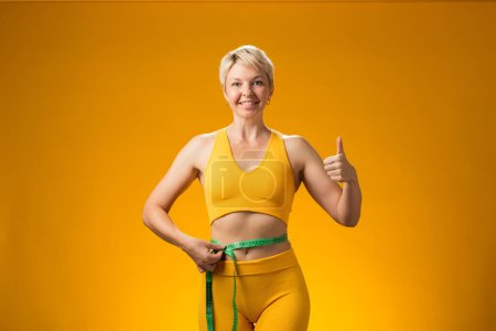 Téléchargez les photos : Femme fitness aux cheveux courts avec ruban à mesurer isolé sur fond jaune. Perte de poids et concept de santé - en image libre de droit