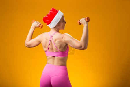 Foto de Mujer con sombrero de Navidad sosteniendo pesas aisladas sobre fondo amarillo. Concepto de vacaciones y fitness - Imagen libre de derechos