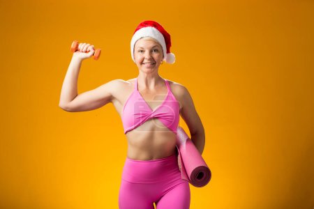 Foto de Sombrero de mujer en Navidad con mancuerna y esterilla de yoga aislada sobre fondo amarillo. Concepto de vacaciones y fitness - Imagen libre de derechos