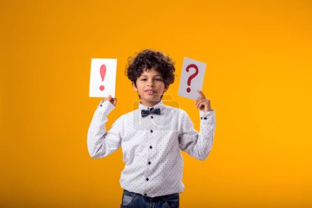 Téléchargez les photos : Portrait d'un enfant garçon tenant des cartes de point d'interrogation et d'exclamation sur fond jaune. Brainstorming et concept de choix - en image libre de droit