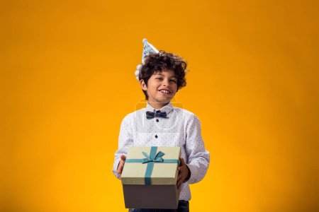 Photo for Portrait of child boy with birthday cap on head holding giftbox over yellow background. Birthday and celebration concept - Royalty Free Image