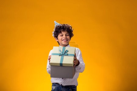 Téléchargez les photos : Portrait d'enfant garçon avec bonnet d'anniversaire sur la tête tenant boîte cadeau sur fond jaune. Concept d'anniversaire et de célébration - en image libre de droit
