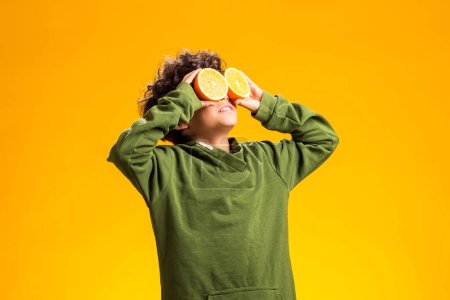Foto de Retrato de niño sosteniendo la mitad de los frutos naranjas frente a los ojos sobre fondo amarillo. Concepto de comida saludable - Imagen libre de derechos