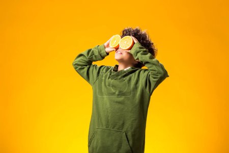 Téléchargez les photos : Portrait d'un enfant garçon tenant la moitié des fruits orange devant les yeux sur fond jaune. Concept d'alimentation saine - en image libre de droit