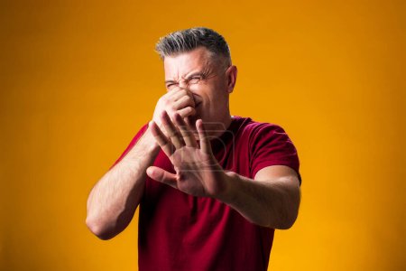 Photo for Portrait of man showing disgust and covering his nose with hand on yellow isolated background, guy refuses bad smell and rejects stench - Royalty Free Image
