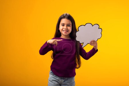 Photo for Portrait of smiling kid girl holding cloud bubble card and pointing finger at it over yellow background. Dreaming concept - Royalty Free Image