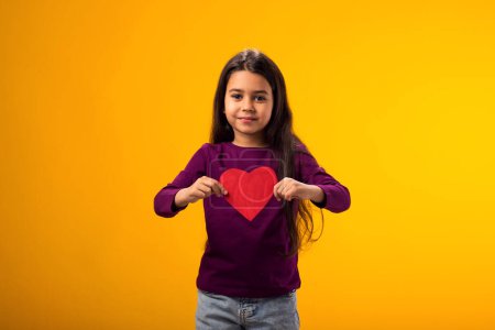 Téléchargez les photos : Portrait d'une enfant fille tenant papier heartover fond jaune. Amour, soins et concept de Saint Valentin - en image libre de droit