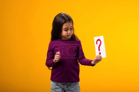Téléchargez les photos : Un portrait d'une gamine réfléchie tenant une carte de point d'interrogation. Enfants, idée et concept de connaissance - en image libre de droit