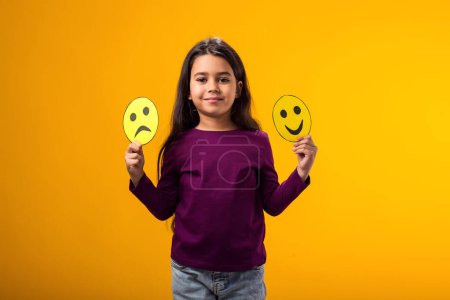 Téléchargez les photos : Portrait de jeune fille souriante tenant émoticônes tristes et heureux dans les mains. Santé mentale, psychologie et émotions des enfants concept - en image libre de droit