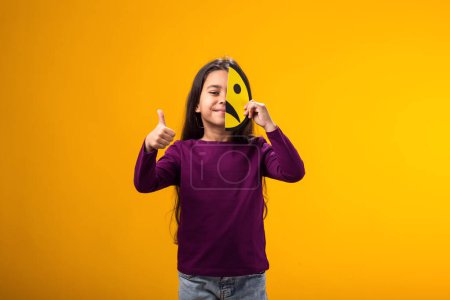 Foto de Retrato de una niña sonriente sosteniendo un triste emoticono de media cara y mostrando un gesto de pulgar hacia arriba. Salud mental, psicología y emociones infantiles - Imagen libre de derechos