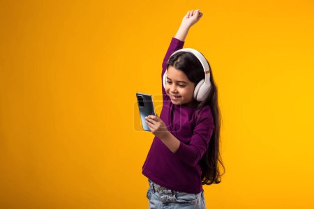 Photo for Portrait of smiling girl enjoying music in headphonesand showing winner gesture on yellow background. Lifestyle, leasure and gadget addiction concept - Royalty Free Image