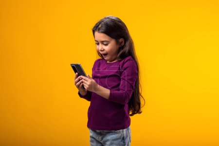 Téléchargez les photos : Portrait d'une petite fille textant un message dans les médias sociaux sur un téléphone portable, utilisant les services de réseau mobile, bavardant en ligne. Studio intérieur tourné isolé sur fond jaune. - en image libre de droit