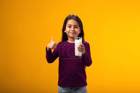 Téléchargez les photos : Portrait d'une enfant souriante tenant un verre de lait et montrant un geste du pouce. Concept nutrition et santé - en image libre de droit