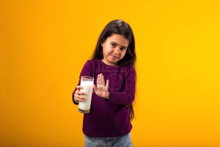 Téléchargez les photos : Portrait d'une fillette allergique aux produits laitiers montrant un geste d'arrêt devant un verre de lait sur fond jaune. Concept d'intolérance au lactose - en image libre de droit