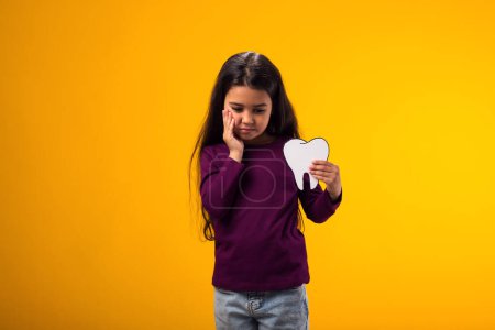 Téléchargez les photos : Portrait d'une fille tenant une dent en papier et touchant la bouche avec la main avec une expression douloureuse à cause de maux de dents ou de maladies dentaires sur les dents. Concept de dentiste. - en image libre de droit