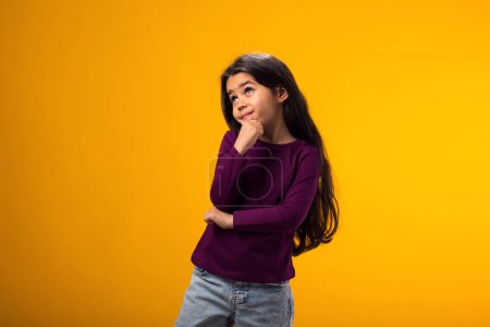 Téléchargez les photos : Portrait d'enfant réfléchi fille toucher le menton avec la pensée du doigt ou de considérer sur fond jaune - en image libre de droit