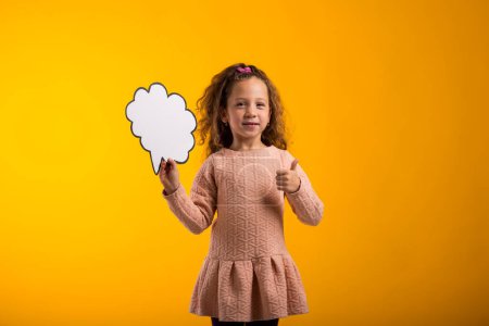 Téléchargez les photos : Portrait d'une jeune fille souriante tenant une carte à bulles et montrant un geste du pouce sur fond jaune. Concept de rêve - en image libre de droit