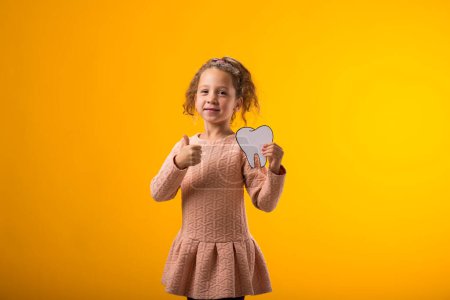 Photo for Portrait of child girl holding papercraft tooth and showing thumb up gesture over yellow background. Dental health concept - Royalty Free Image