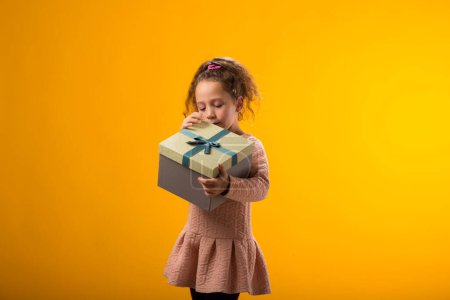 Photo for Portrait of smiling kid girl holding giftbox and looking inside the box over yellow background. Birthday and celebration concept - Royalty Free Image