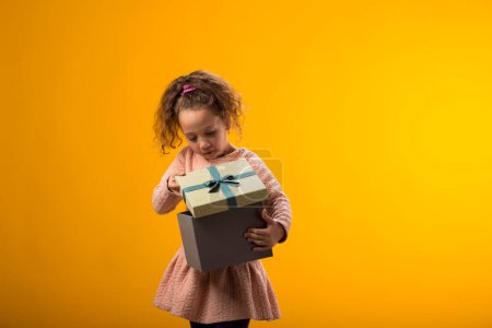 Photo for Portrait of smiling kid girl holding giftbox and looking inside the box over yellow background. Birthday and celebration concept - Royalty Free Image